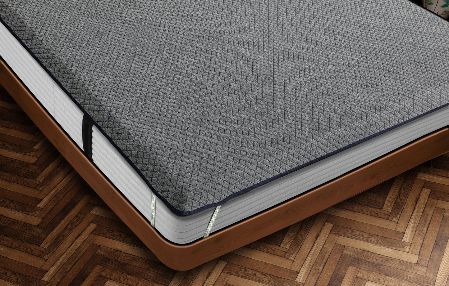 Close-up corner view of Durfi Essential Mattress Protector with grey diamond-quilted fabric and navy piping fitted snugly over a mattress on a warm brown wooden bed frame with herringbone parquet flooring