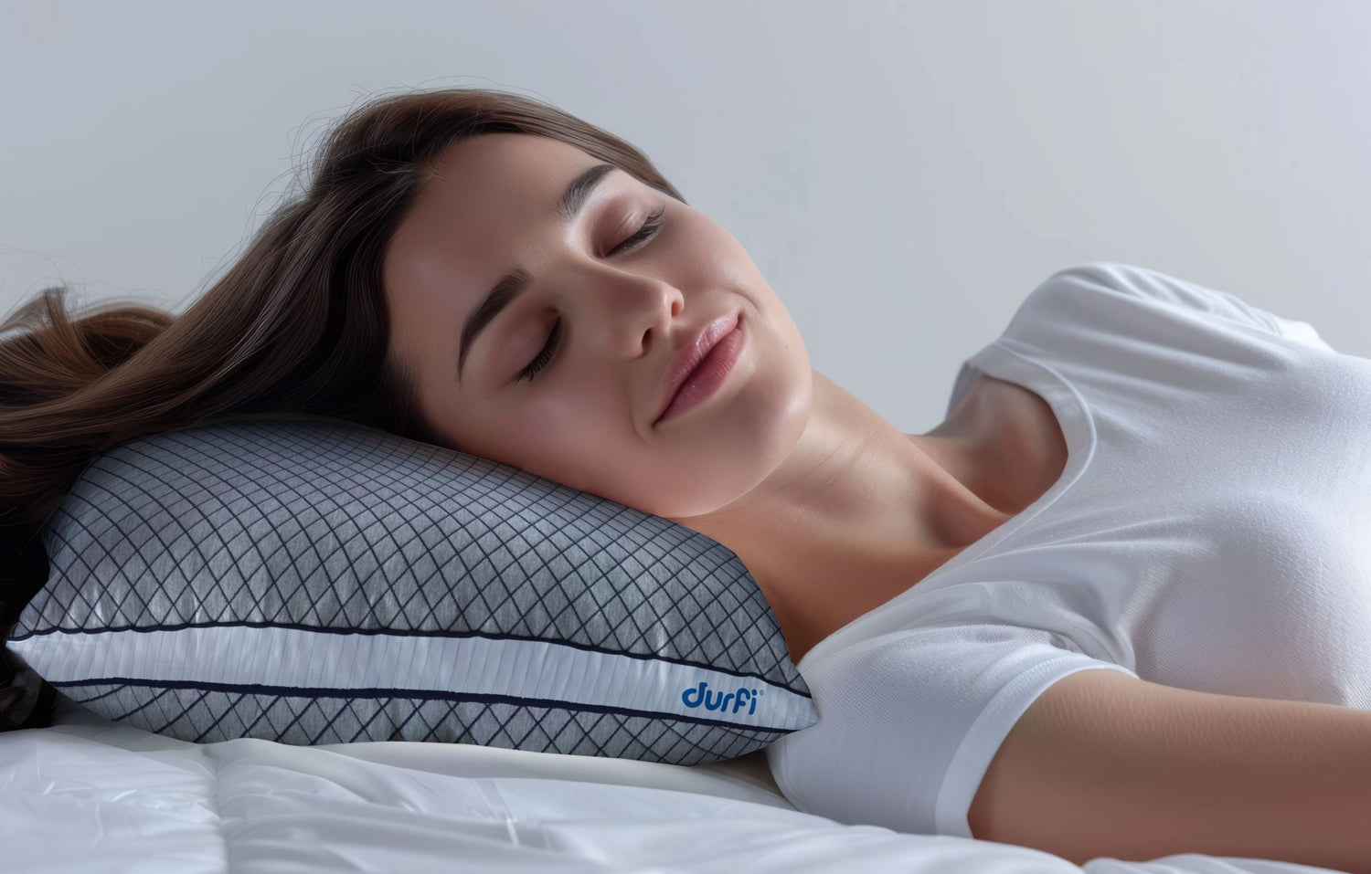 Woman sleeping comfortably on her side on a Durfi Dual Comfort Dr. Pillow with grey diamond-quilted cover and navy piping, shown on a white bed in a soft-lit bedroom