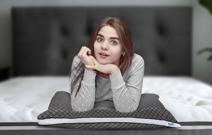 Young woman resting her chin on a Durfi Dual Comfort Pillow while lying on a white mattress in front of a dark tufted headboard, looking cheerfully at the camera