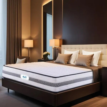 Durfi Hybrid Pocket Spring Mattress shown at an angle on a bed in an upscale hotel-style bedroom with a tall beige headboard, warm lighting, and decorative brown cushions